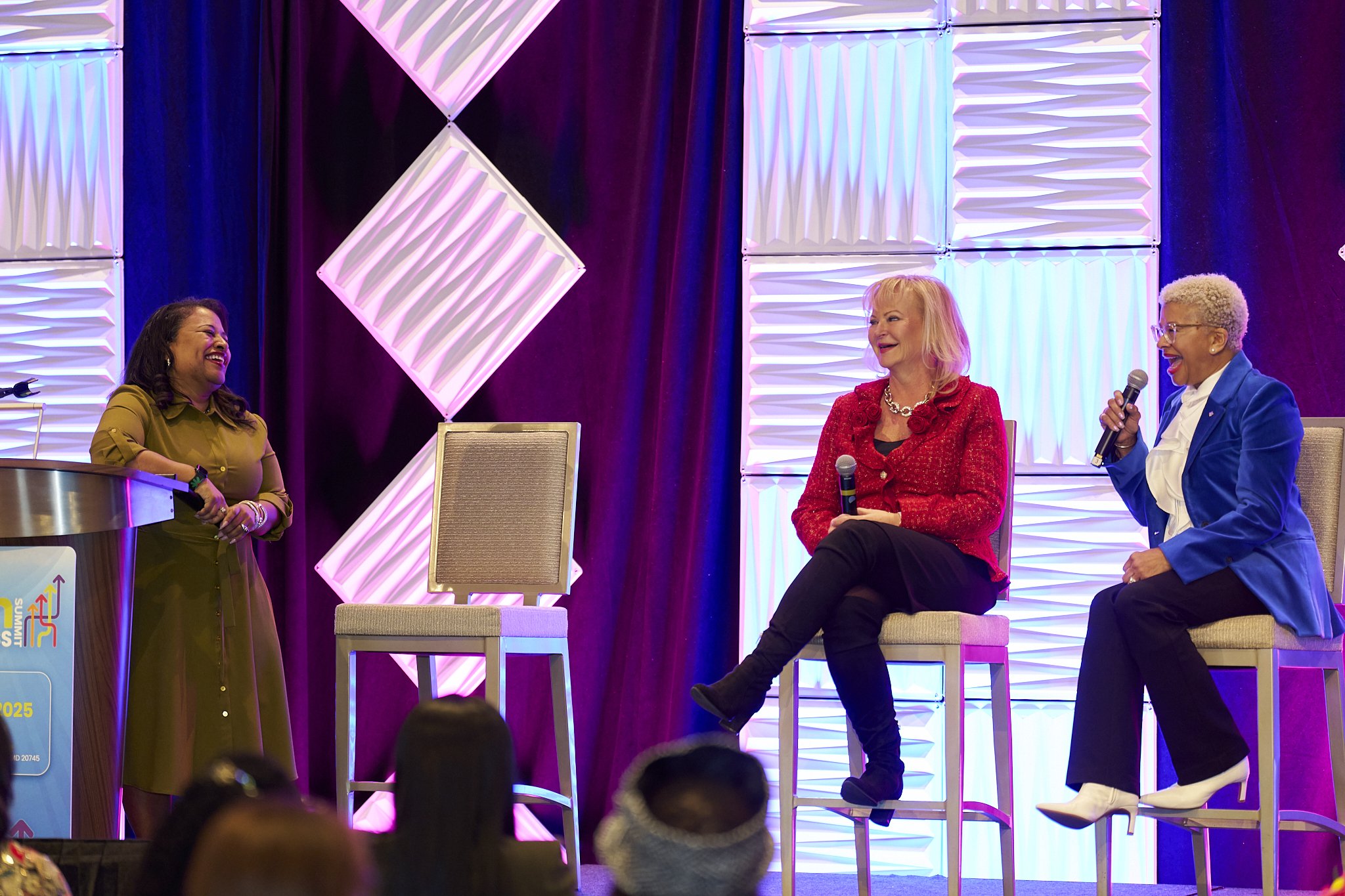Three women on stage at a conference or panel discussion, with two sitting and one standing. The woman on the left is standing near a podium, the two women on the right are seated on high chairs. They are all smiling and holding microphones. The back