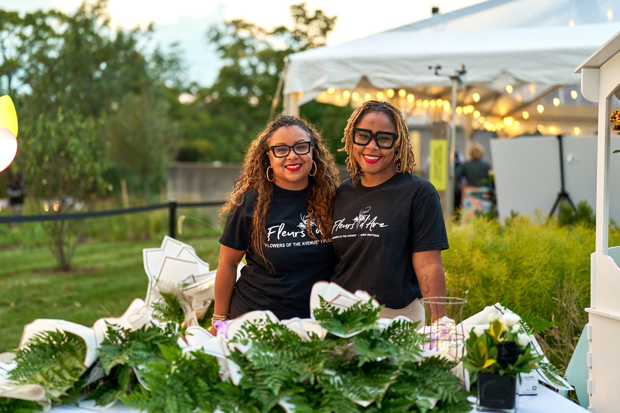 Two smiling women with curly hair and glasses standing behind a table full of flowers at an outdoor event during the evening. Parks and People.