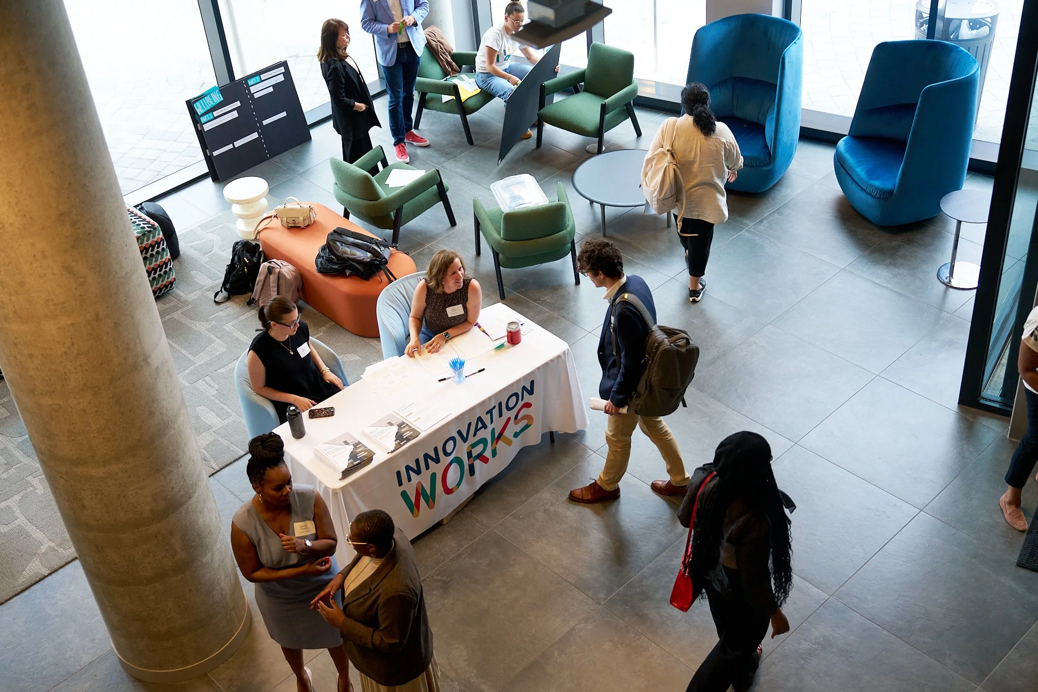 People at a registration or information desk at an event with a banner that reads 'Innovation Works' in a modern lobby with seating areas and large glass windows.