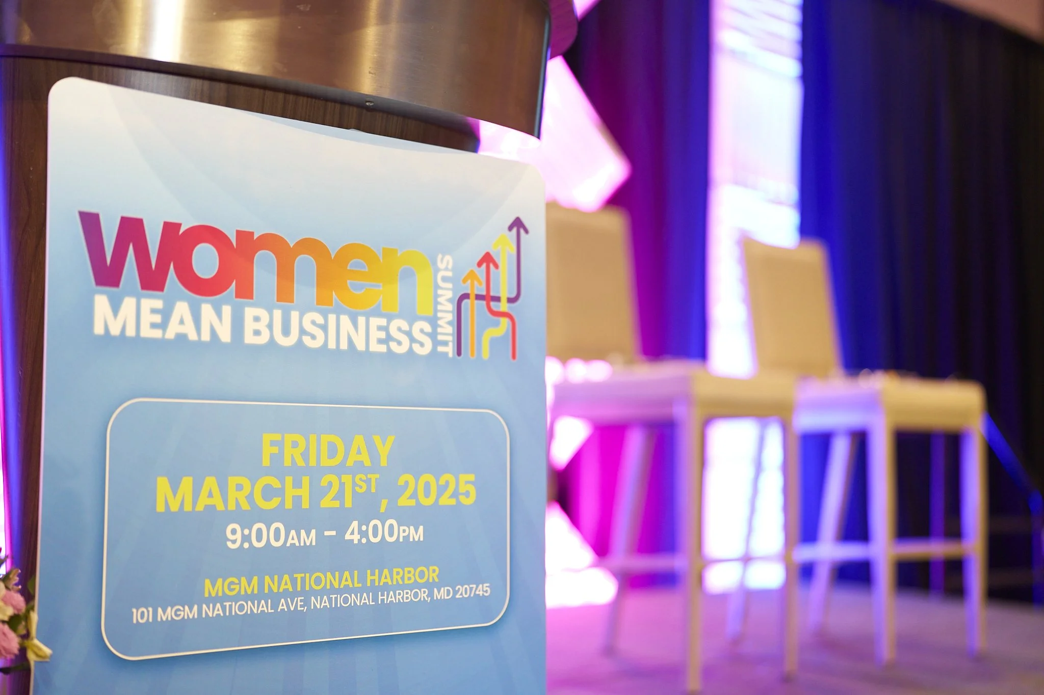 Sign for Women Mean Business Summit on March 21, 2025, at MGM National Harbor, with empty chairs and stage in the background.