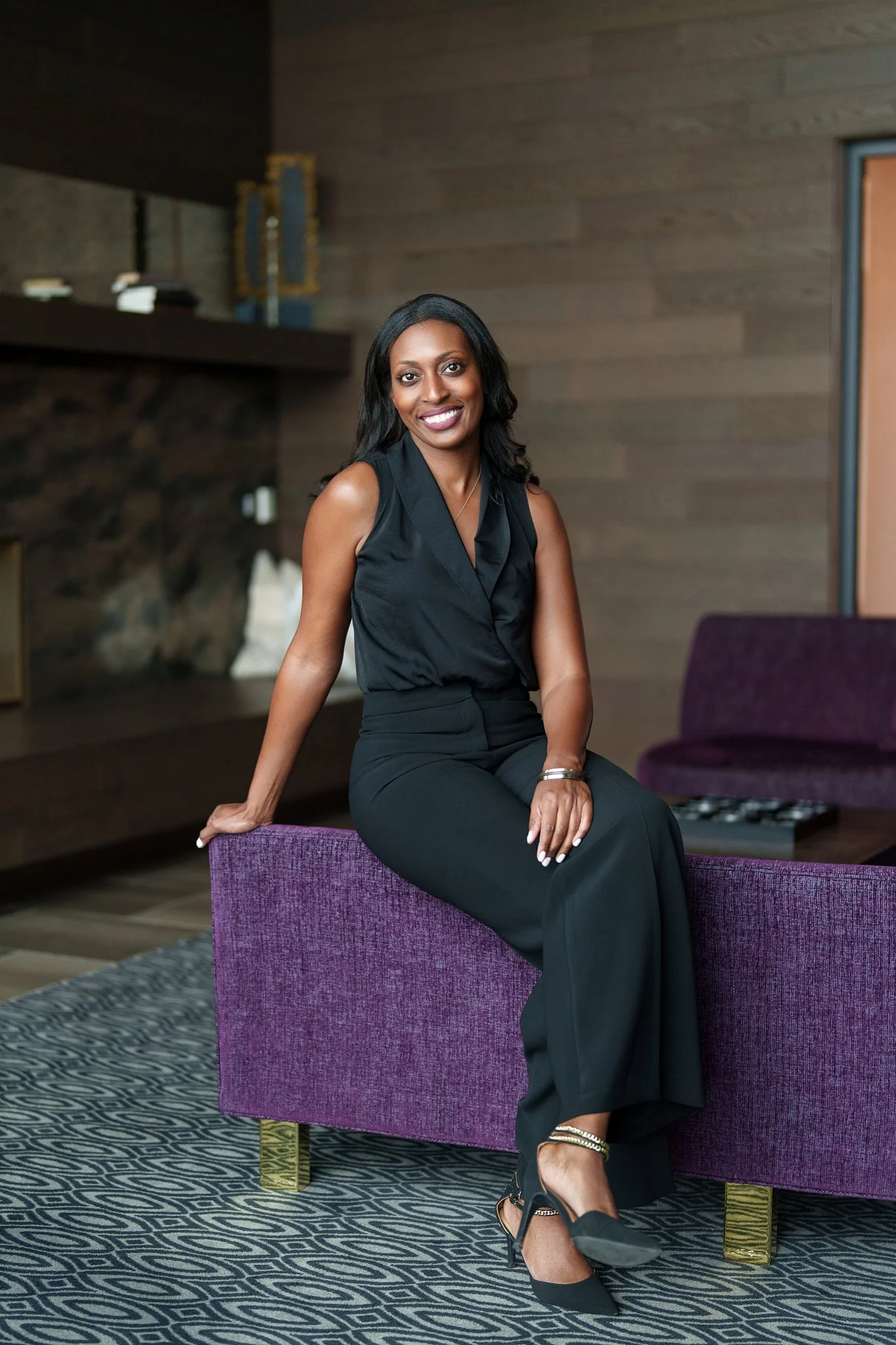 A woman dressed in black, sitting on a purple couch in a modern, wood-paneled room, smiling at the camera. Sophia McCormick. Real Estate Agent.