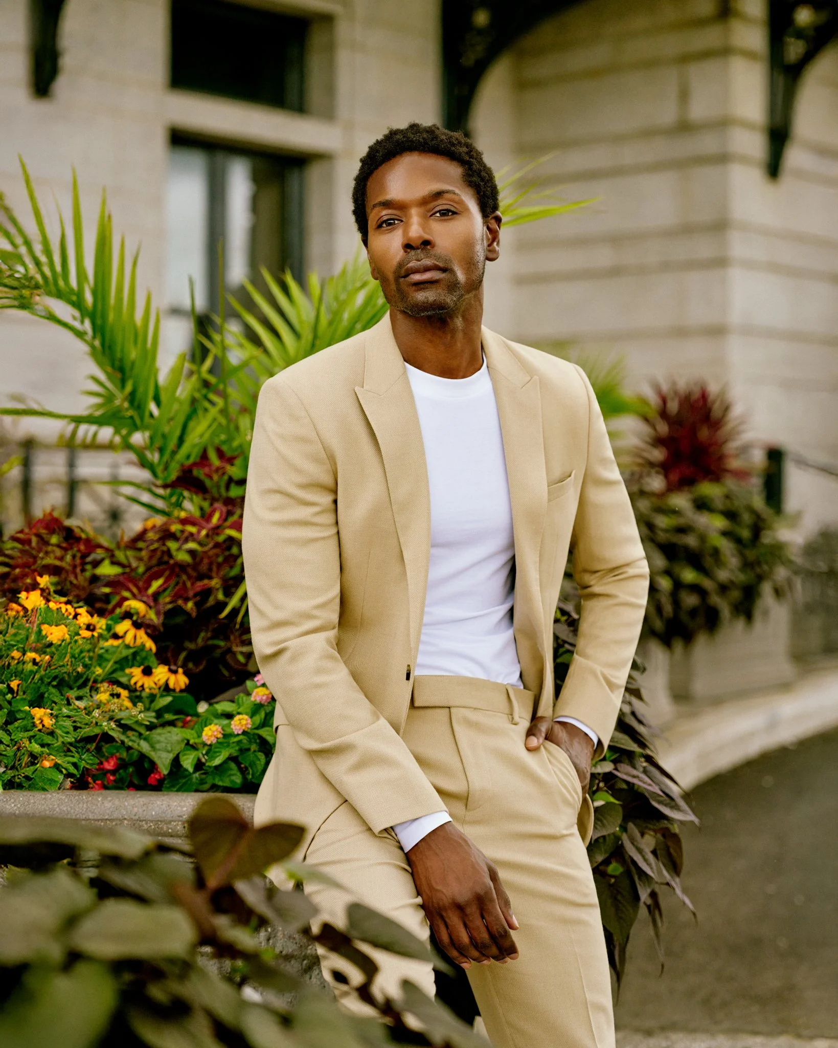 A man in a beige suit and white t-shirt sitting on a stone bench outside surrounded by plants and flowers.
