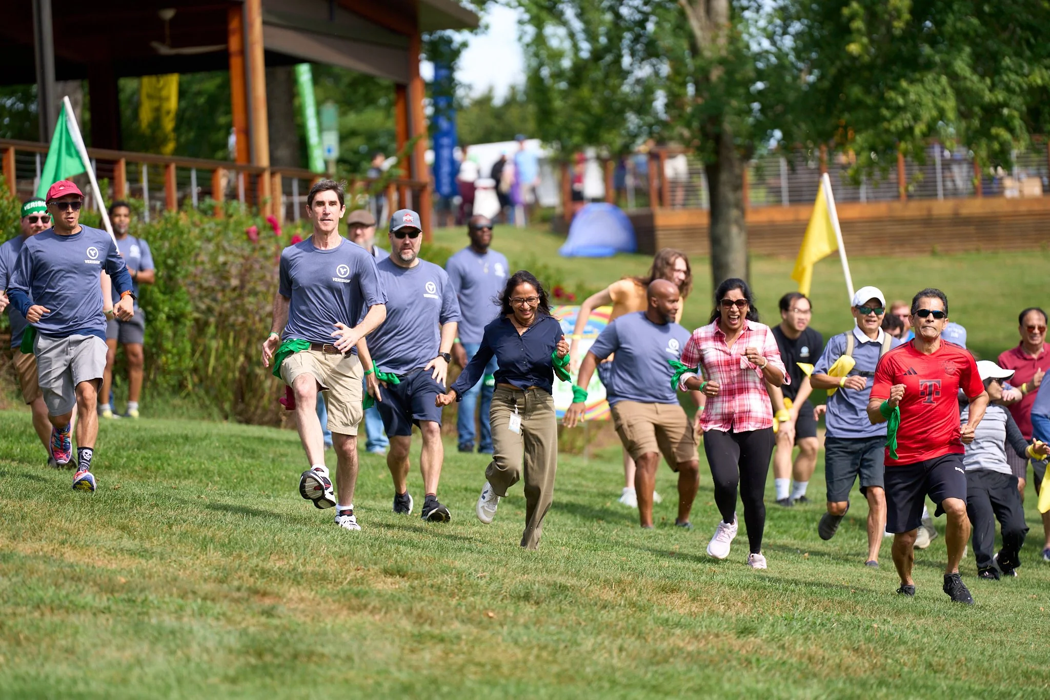 Group of diverse people running outdoors on a grassy field during daytime at an event, with some holding green ribbons and flags, in a park with trees and a wooden structure in the background. VERISIGN