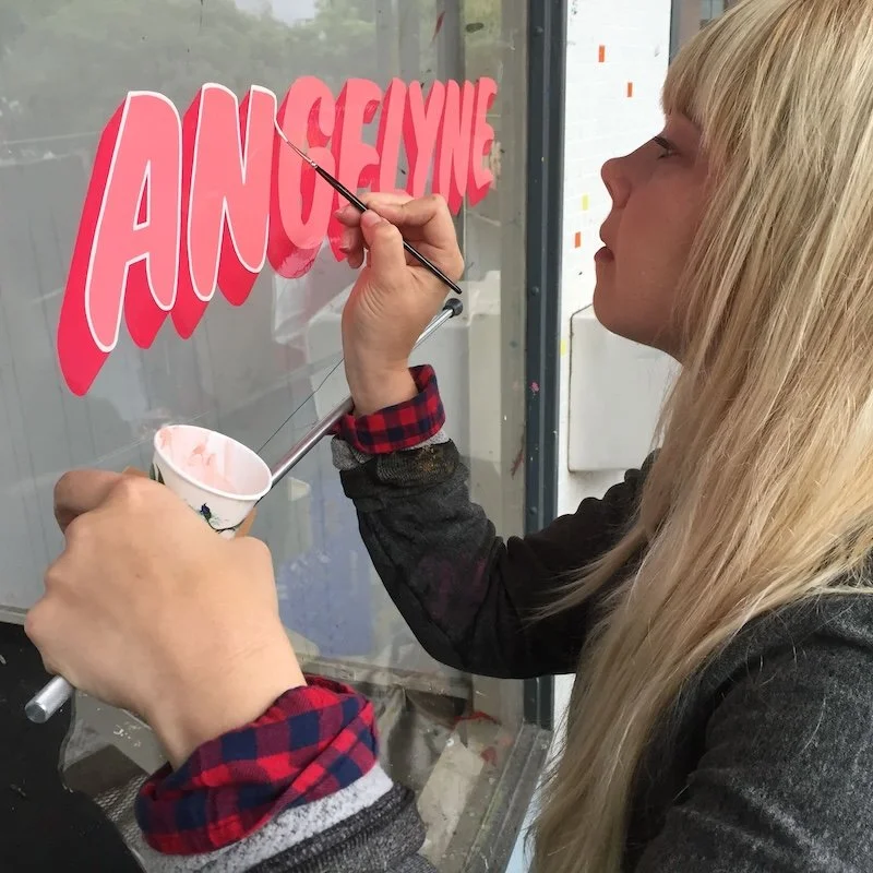 A woman paints a store-front sign on a window, "Angelyne"