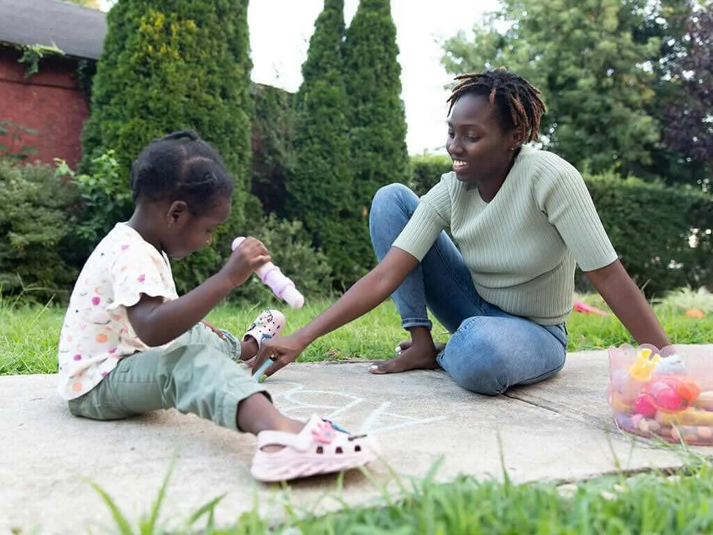 Mother and preschool daughter sitting on outdoor sidewalk doing art activity together with chalk and paint, demonstrating parent-child learning interaction