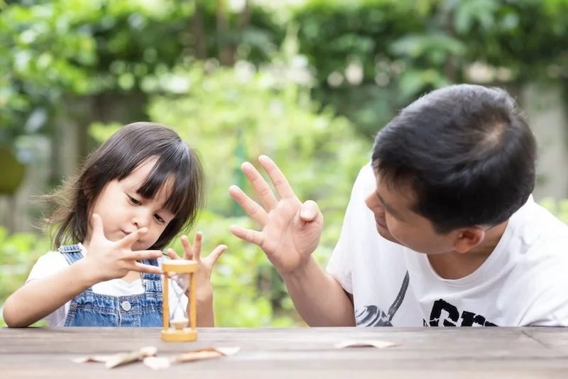 A father and daughter outside at a wooden table practice counting on their fingers with an hourglass on the table.