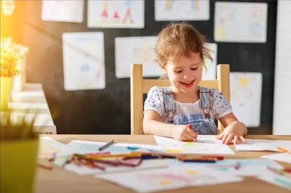 Photo of a smiling preschool girl with bangs at a table in a sunny room coloring art with crayons with blackboard in back decorated with drawings