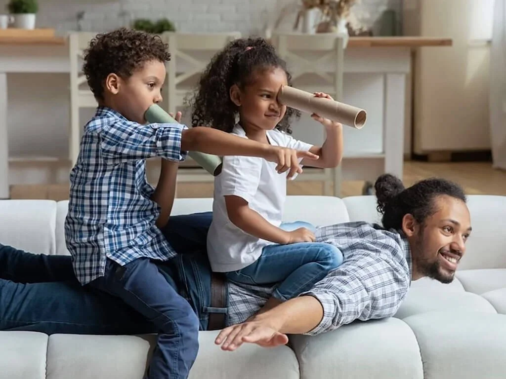 A dad pretends to be an airplane on a couch while two children sit on his back and pretend they are flying and exploring with paper towel rolls