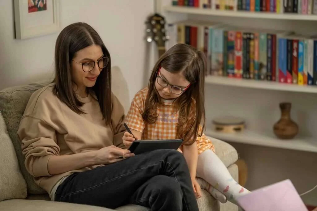Mother and young daughter sitting together reading tablet or book, showing intentional family learning moment and parent-child closeness through shared reading
