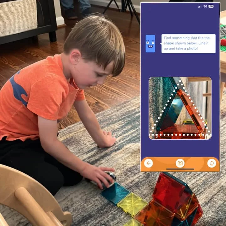 A photo of a 5-year-old boy  looking for triangles and taking pictures of magnet toys shaped like triangles with an overlay image of an app and character talk bubble that says "find something that fits the shape shown below"