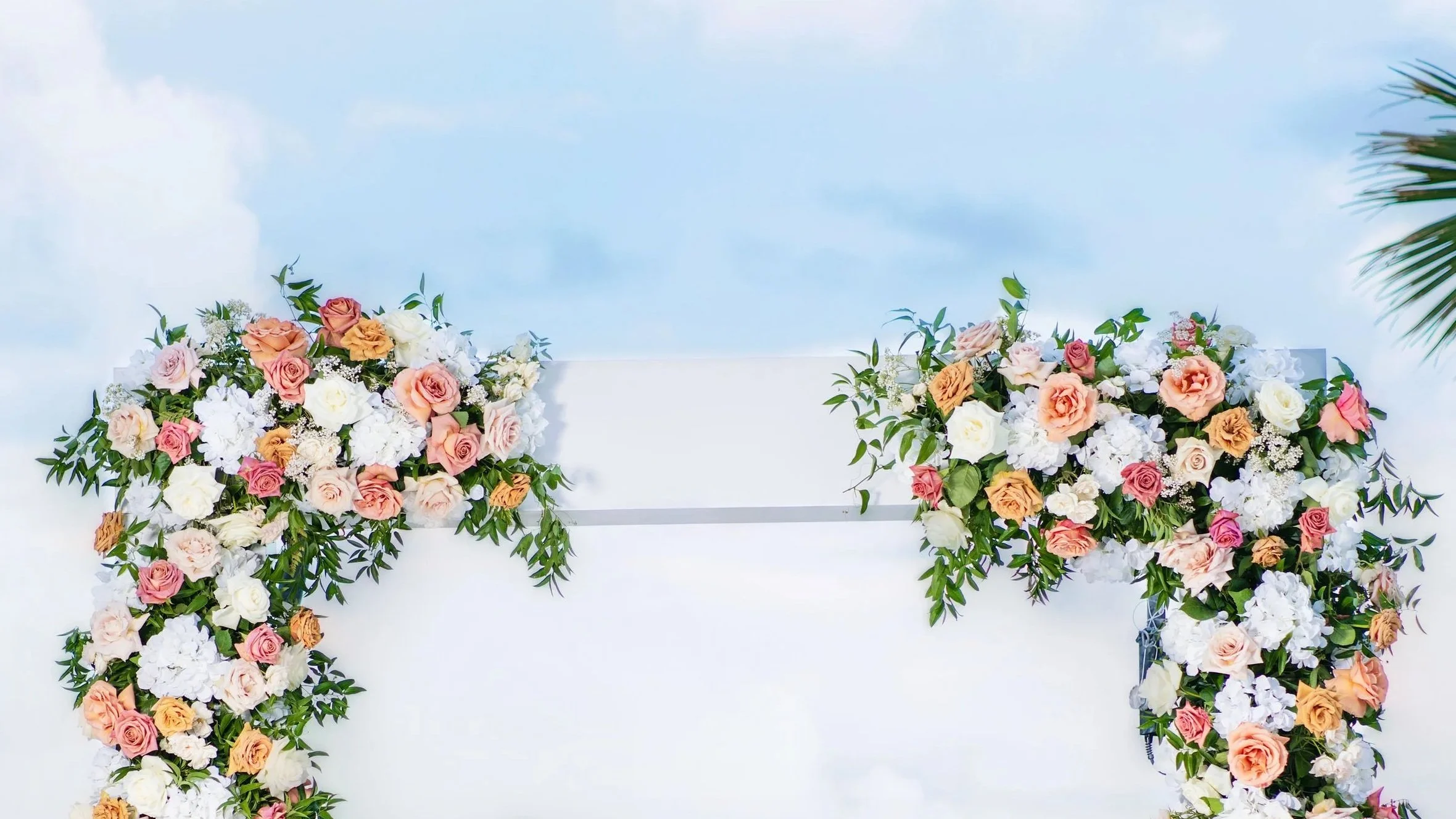 Decorative wedding arch adorned with pink, peach, cream, and white roses along with green foliage, set against a cloudy sky with a palm tree visible on the right.