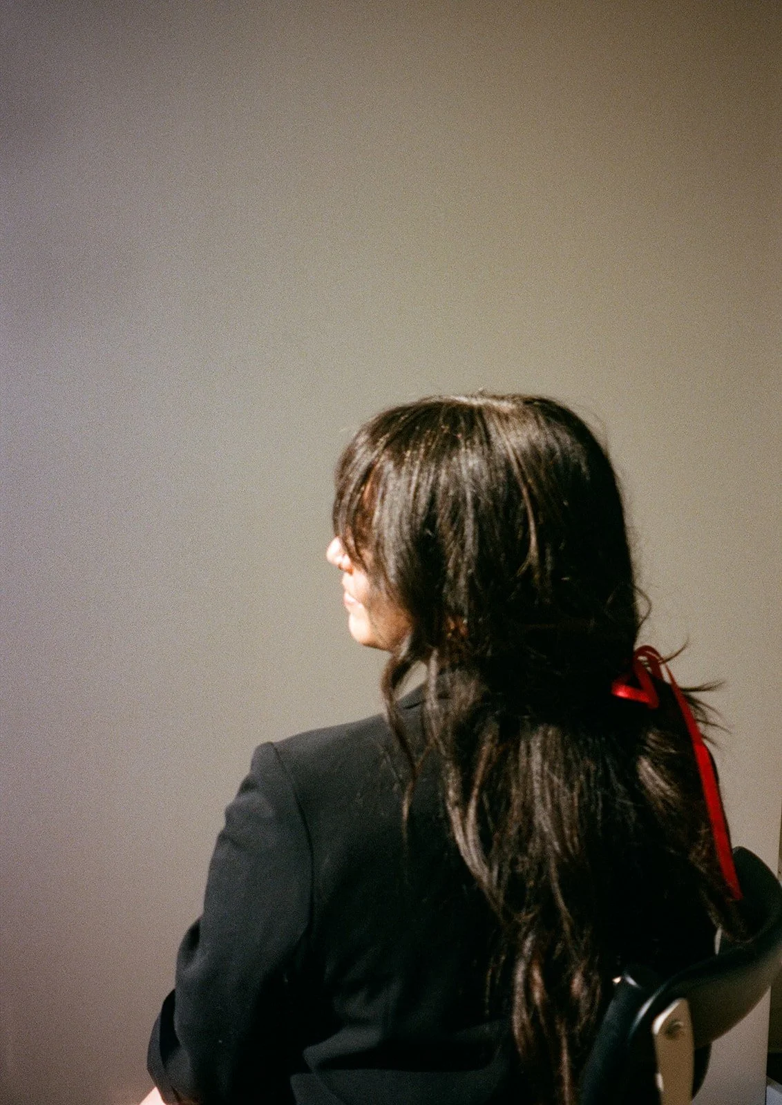 Side view of a woman with dark wavy hair wearing a black blazer, sitting on a chair with a plain neutral background.