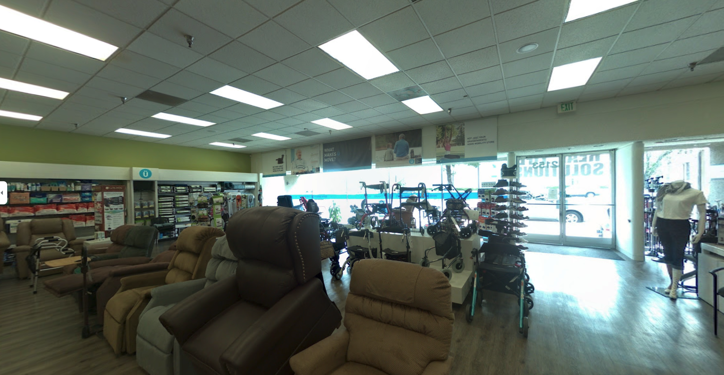 Interior view of a store with various mobility aids like wheelchairs, walkers, and scooters, along with recliner chairs and a mannequin dressed in a white top and black skirt near the front entrance.