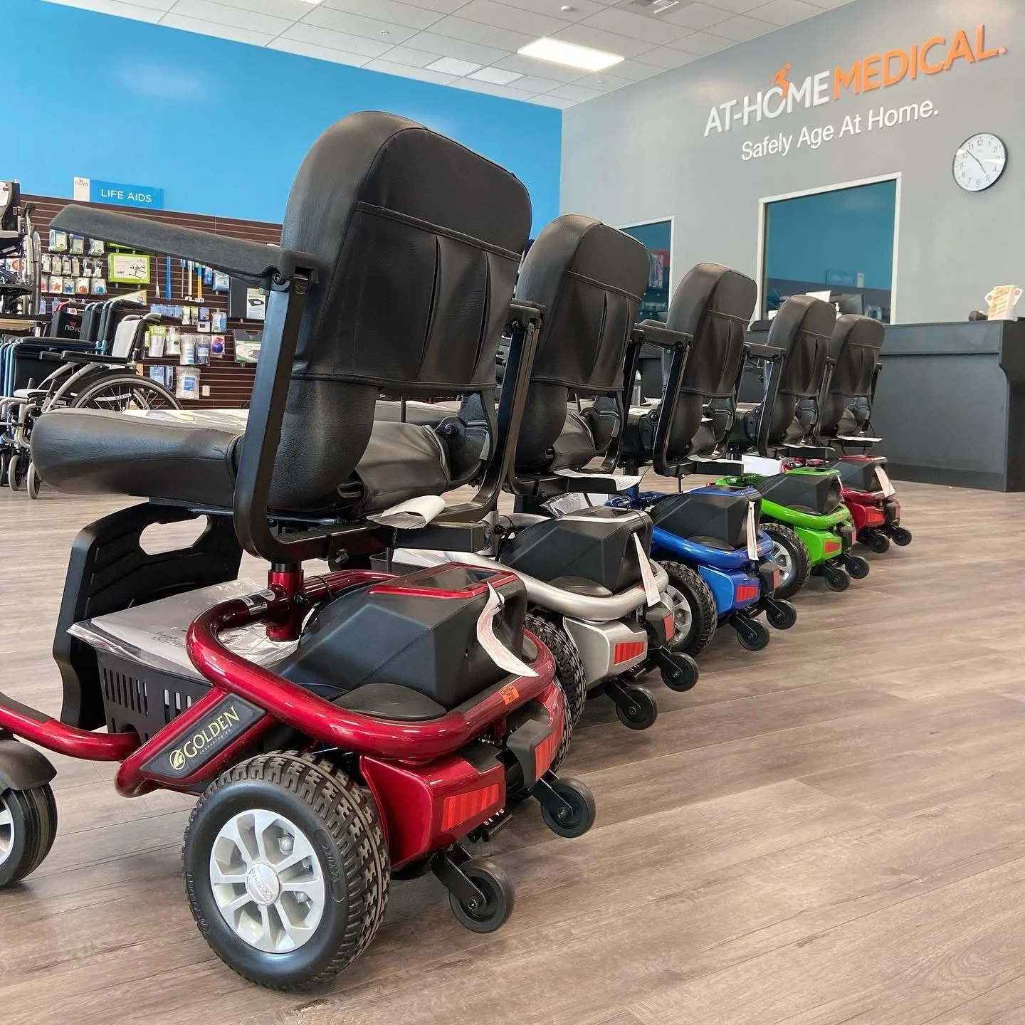 A row of mobility scooters with black seats and colorful bases parked inside a medical supply store.