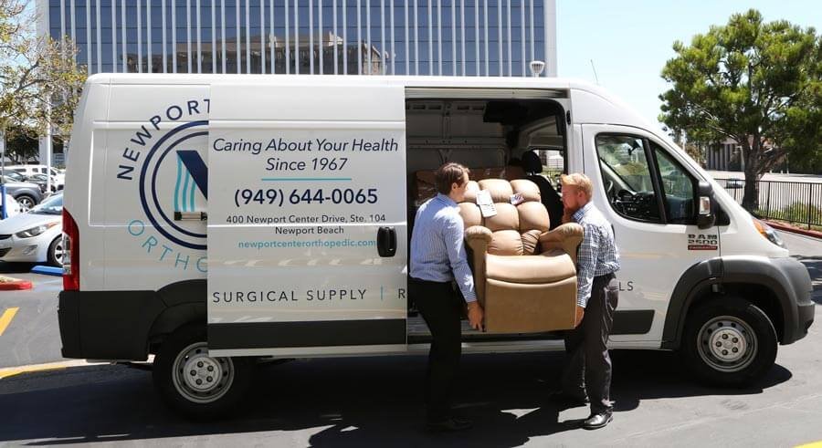 Two men standing outside a white delivery van with the logo and contact information for Newport Orthopedic Surgery. The van's side door is open, revealing a large piece of medical equipment or furniture being loaded or unloaded.