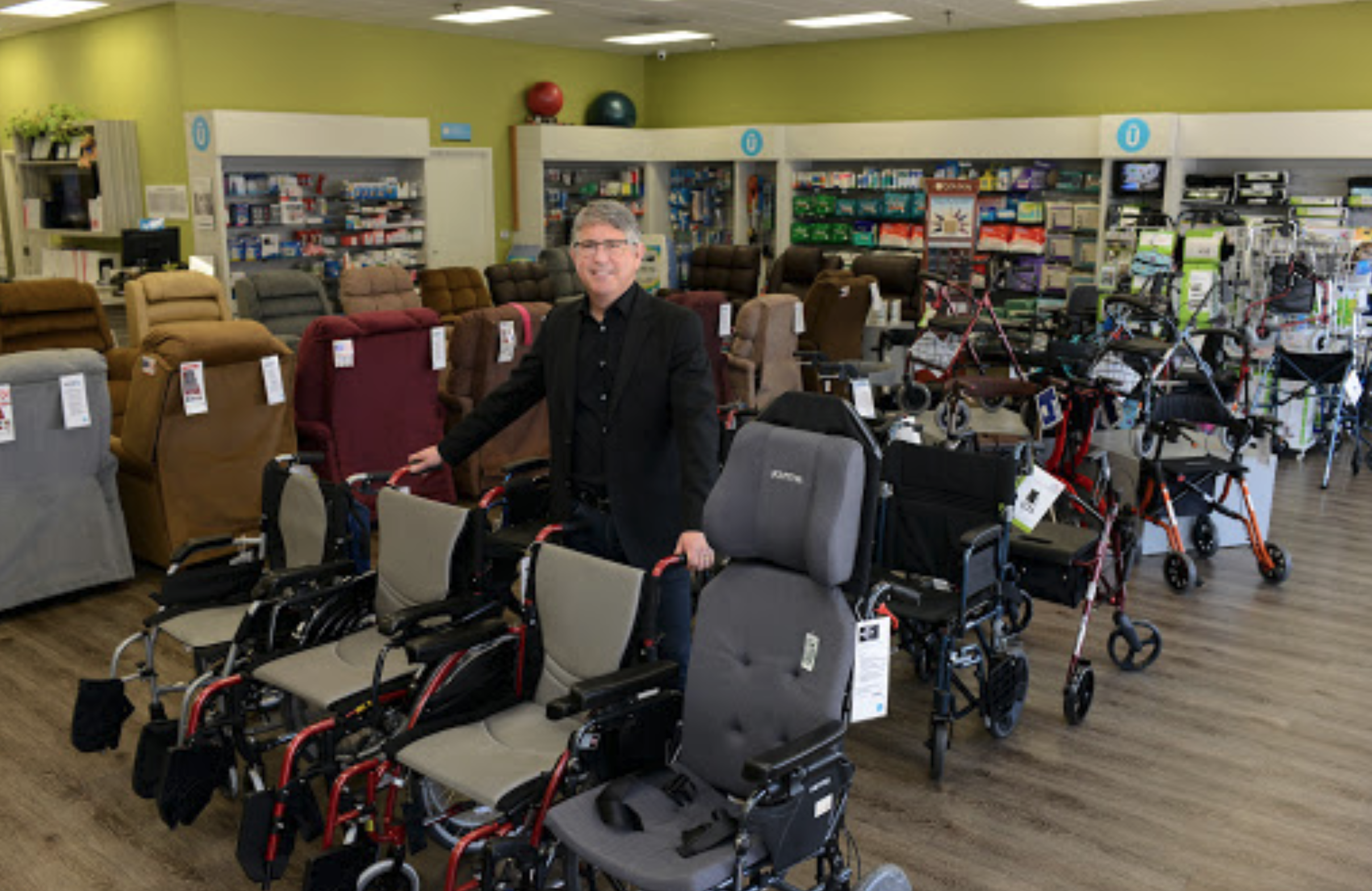 Man in black suit shopping for mobility aids and wheelchairs at a store with various chairs, walkers, and medical supplies on shelves.