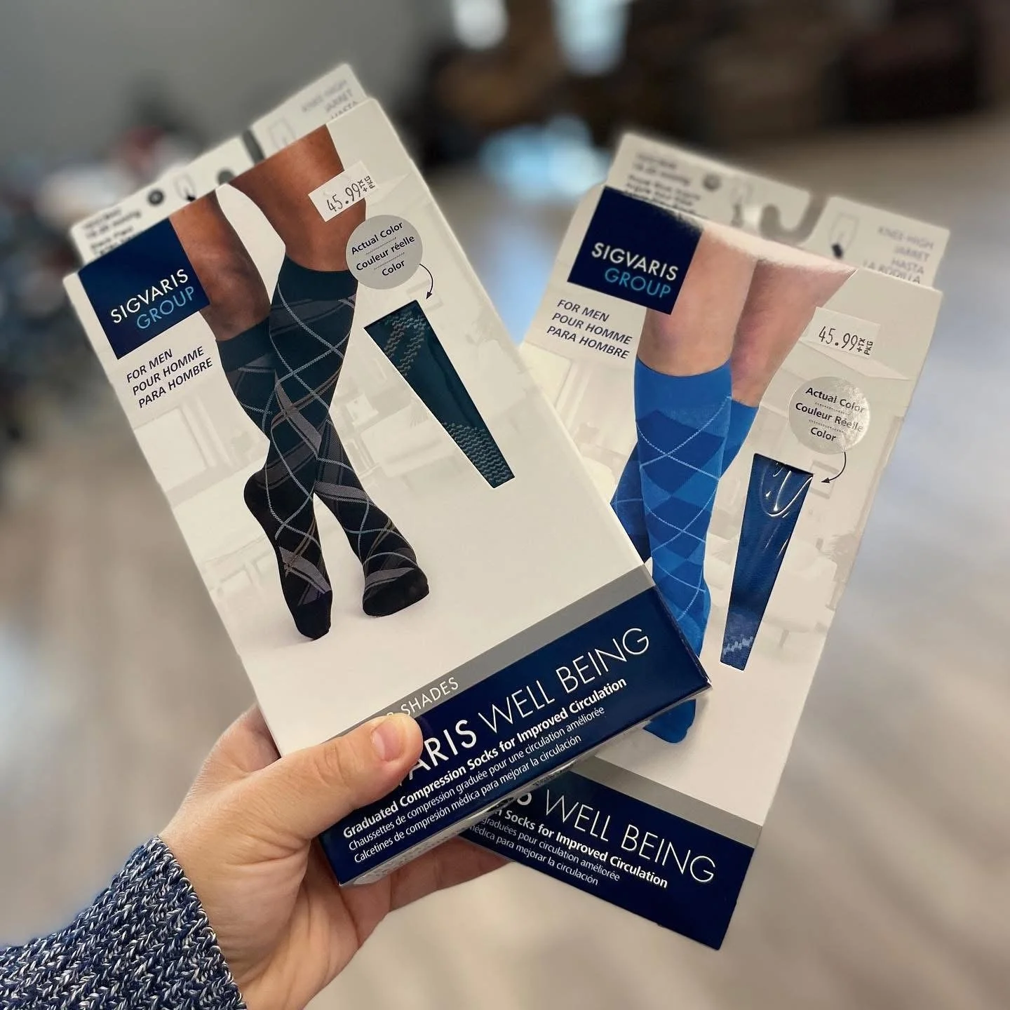 Two boxes of men's compression socks with argyle pattern packaging, one black with plaid pattern and the other blue with diamond pattern, held by a person wearing a blue patterned sleeve.
