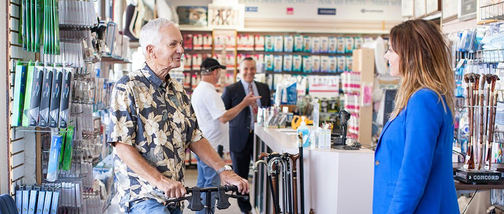 A man with a walker speaking to a woman behind a checkout counter in a store. The store has various items on shelves, and another customer in the background.