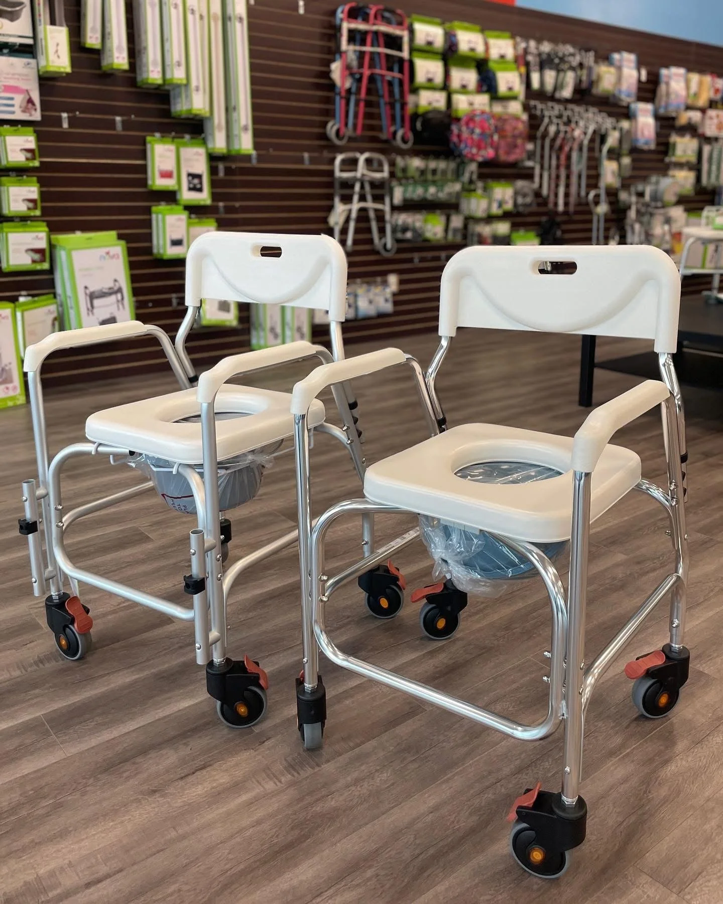 Two white shower chairs on wheels with armrests and backrests displayed in a store aisle, with various mobility aids and accessories on the wall behind them.