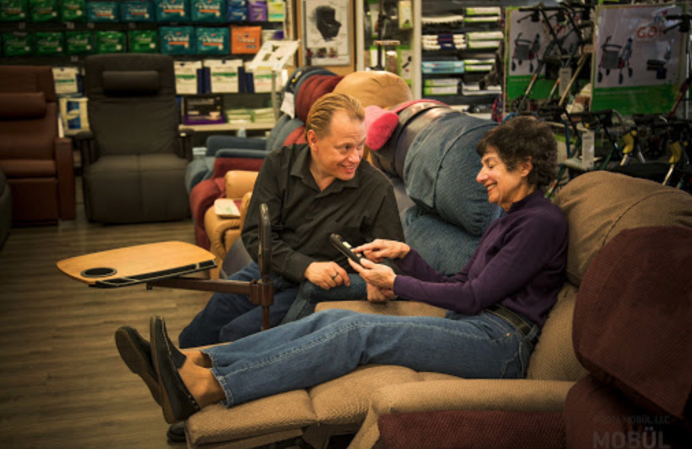Two people sitting on a sofa in an electronics store, smiling and talking, with shelves of chairs, walker carts, and boxed items in the background.