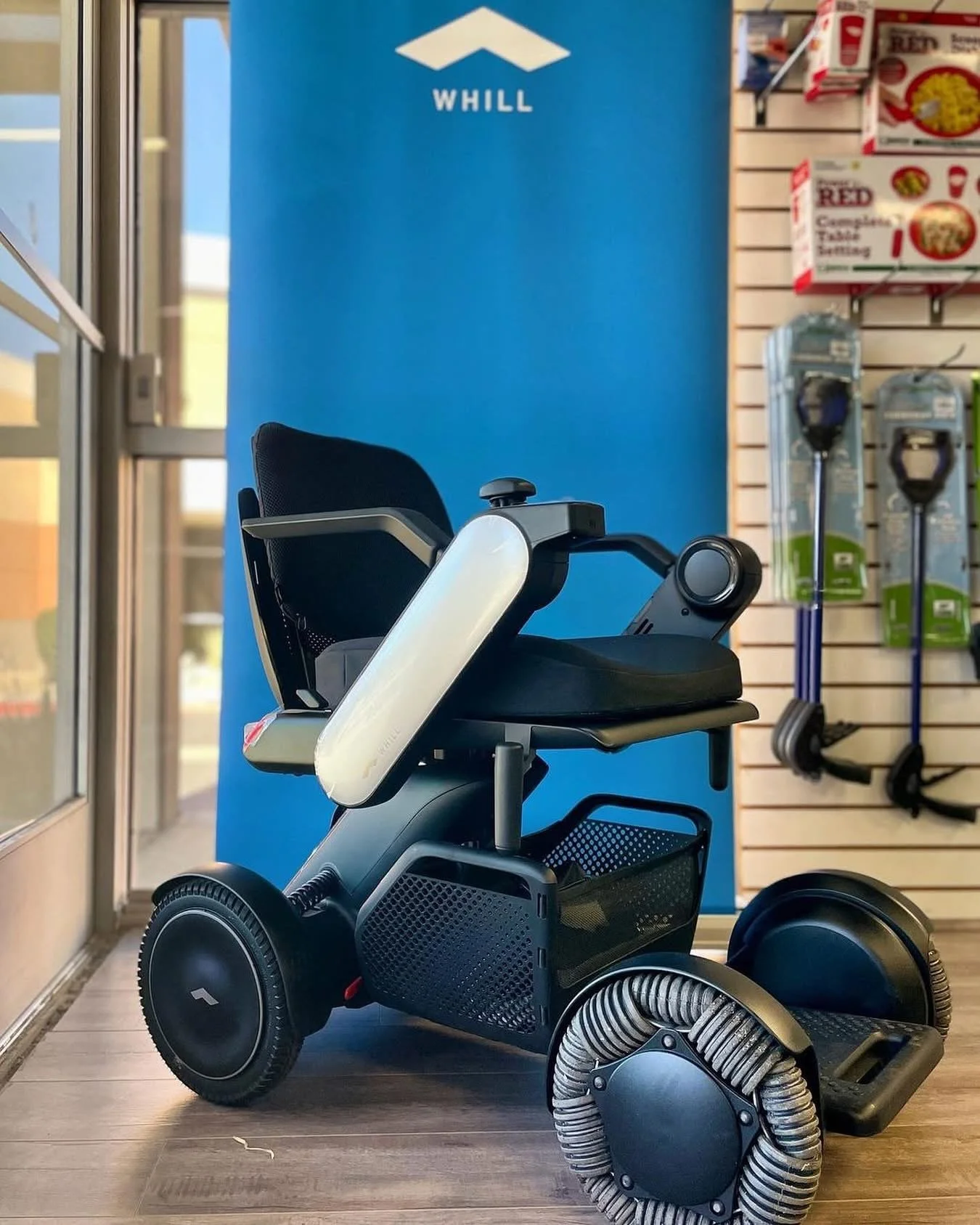 A mobility scooter with large rear wheels and a seat, positioned inside a store near a blue wall with the word 'WHILL' on it. In the background, there are cleaning tools hanging on a wall.