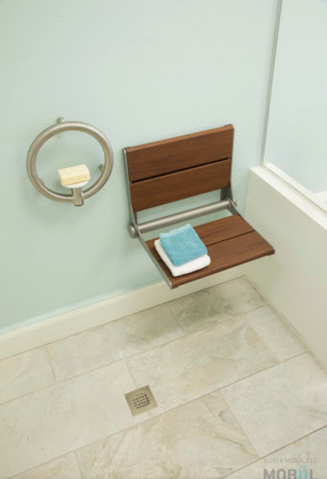A shower bench with folded blue and white towels in a bathroom corner, with a soap dish attached to the wall and a floor drain.