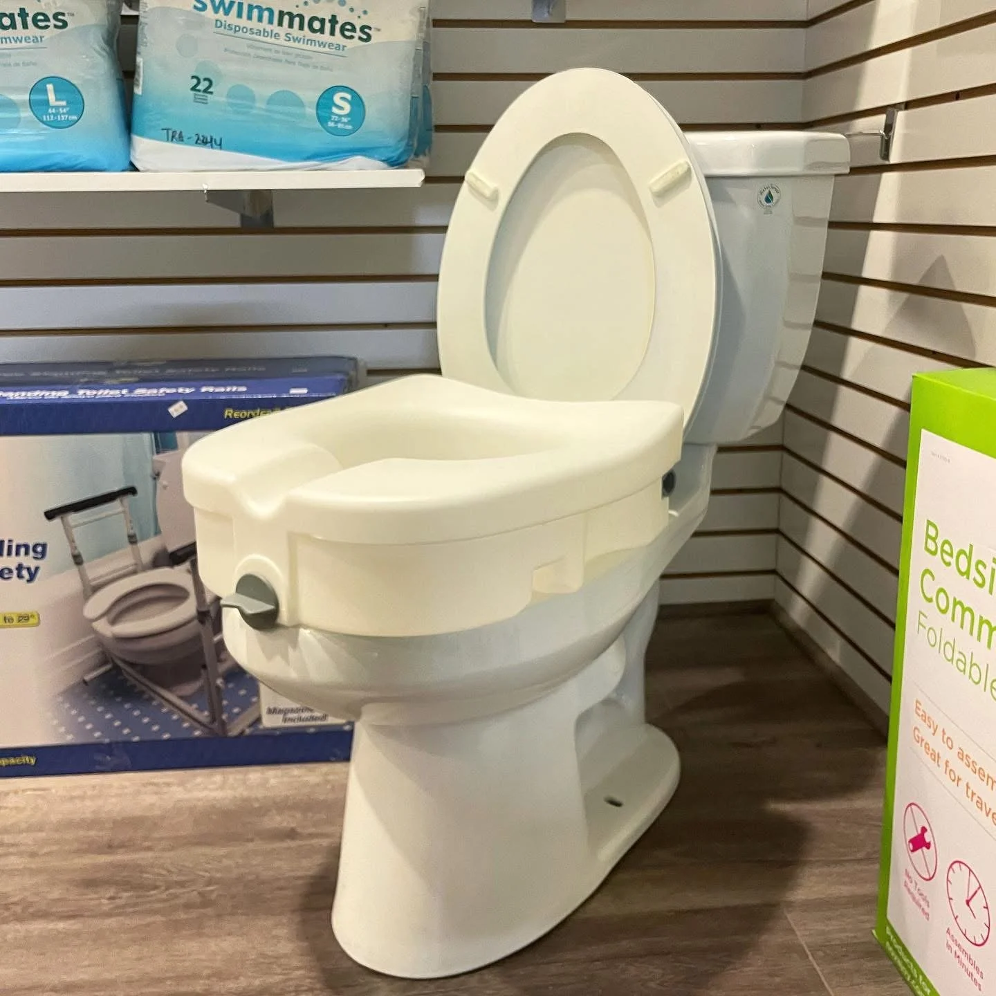 A white portable toilet with a raised lid, displayed on a store shelf among other medical and health products.
