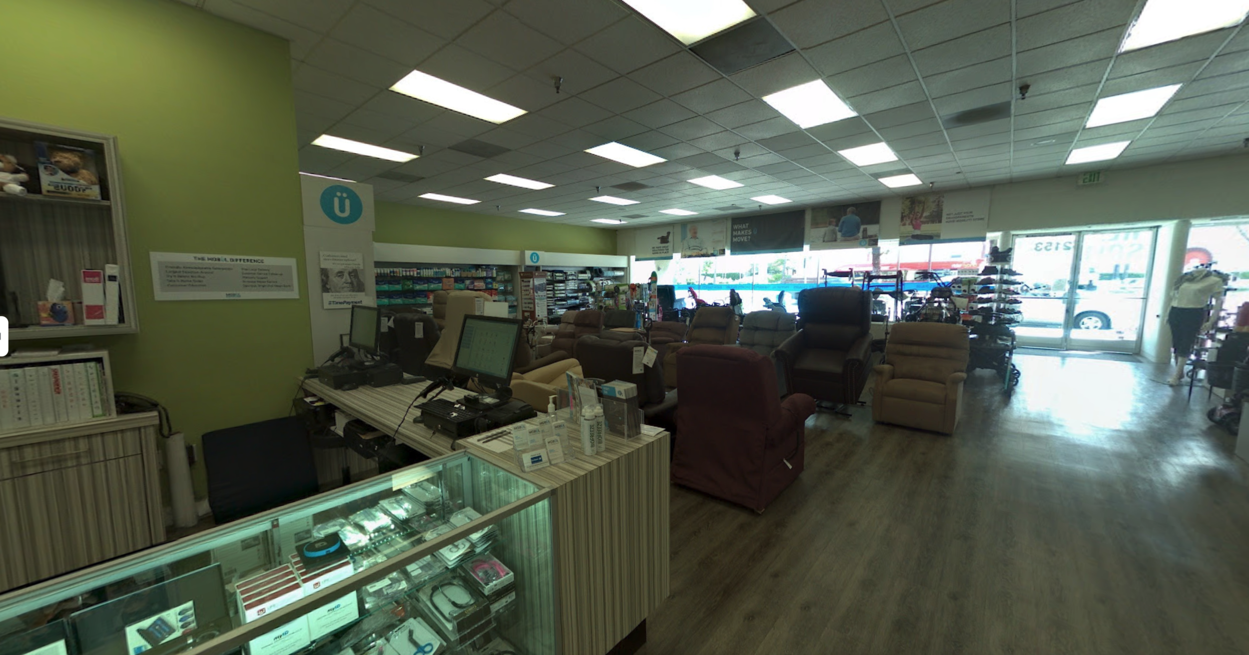 Interior of a furniture store with various recliners and chairs on display near the window, and checkout counters with computers. To the right, shelves with accessories and a person walking near the entrance.