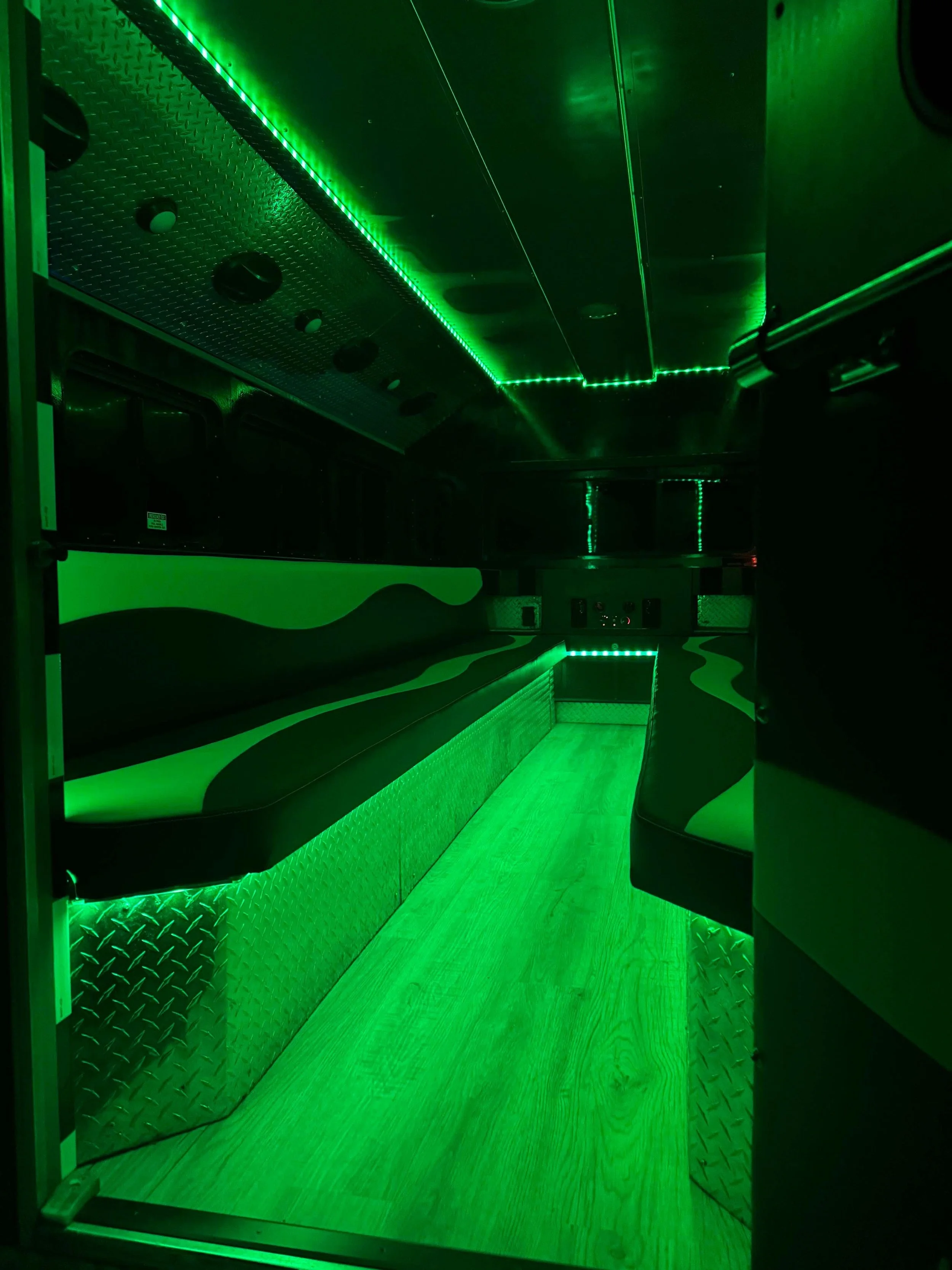 Interior of a vehicle with green LED lighting, equipped with cushioned seating along the sides, and wooden flooring.