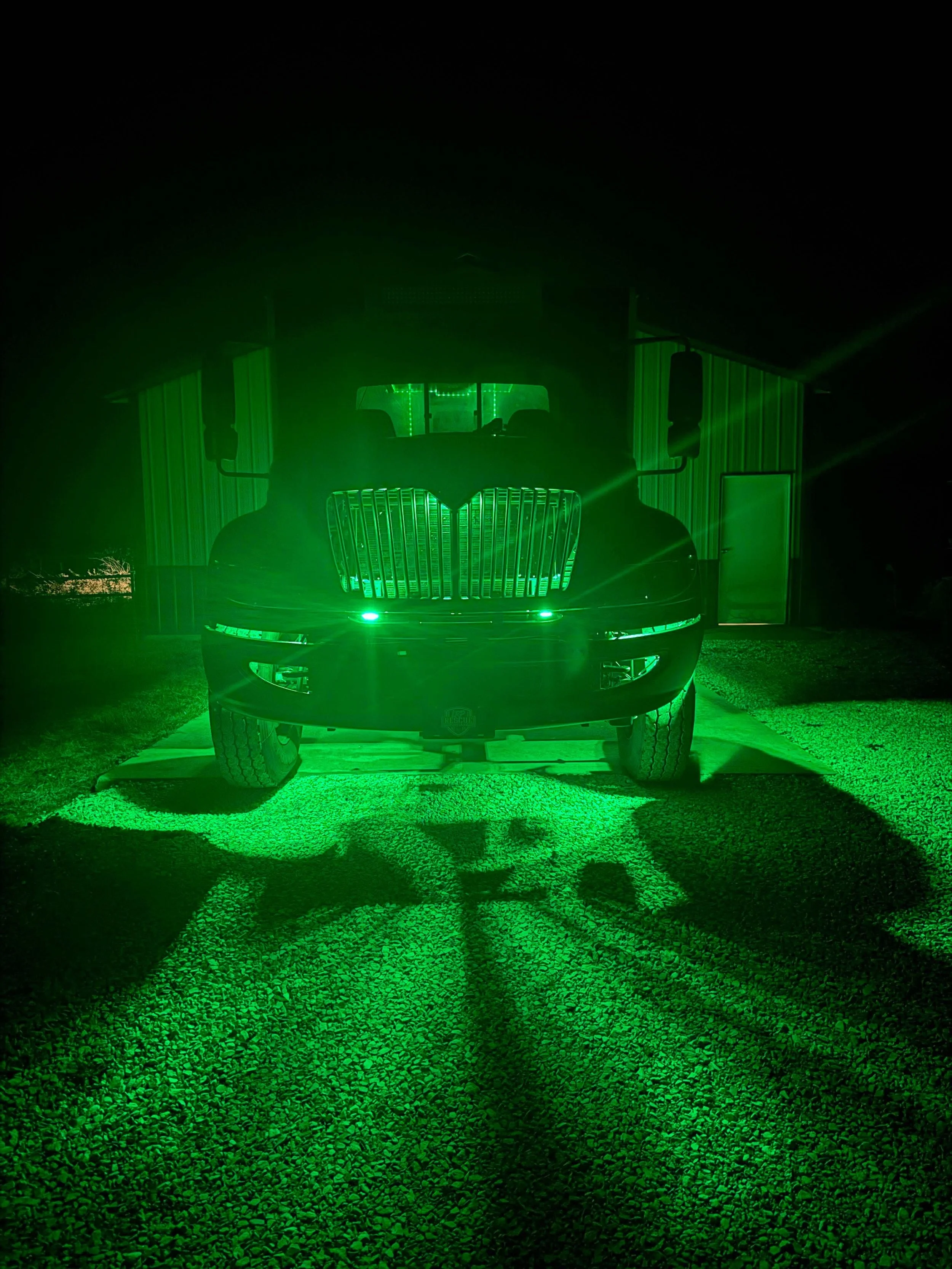 A large truck illuminated by green lights outside a building at night. The front grille and headlights are visible, and the truck casts a green shadow on the gravel ground.