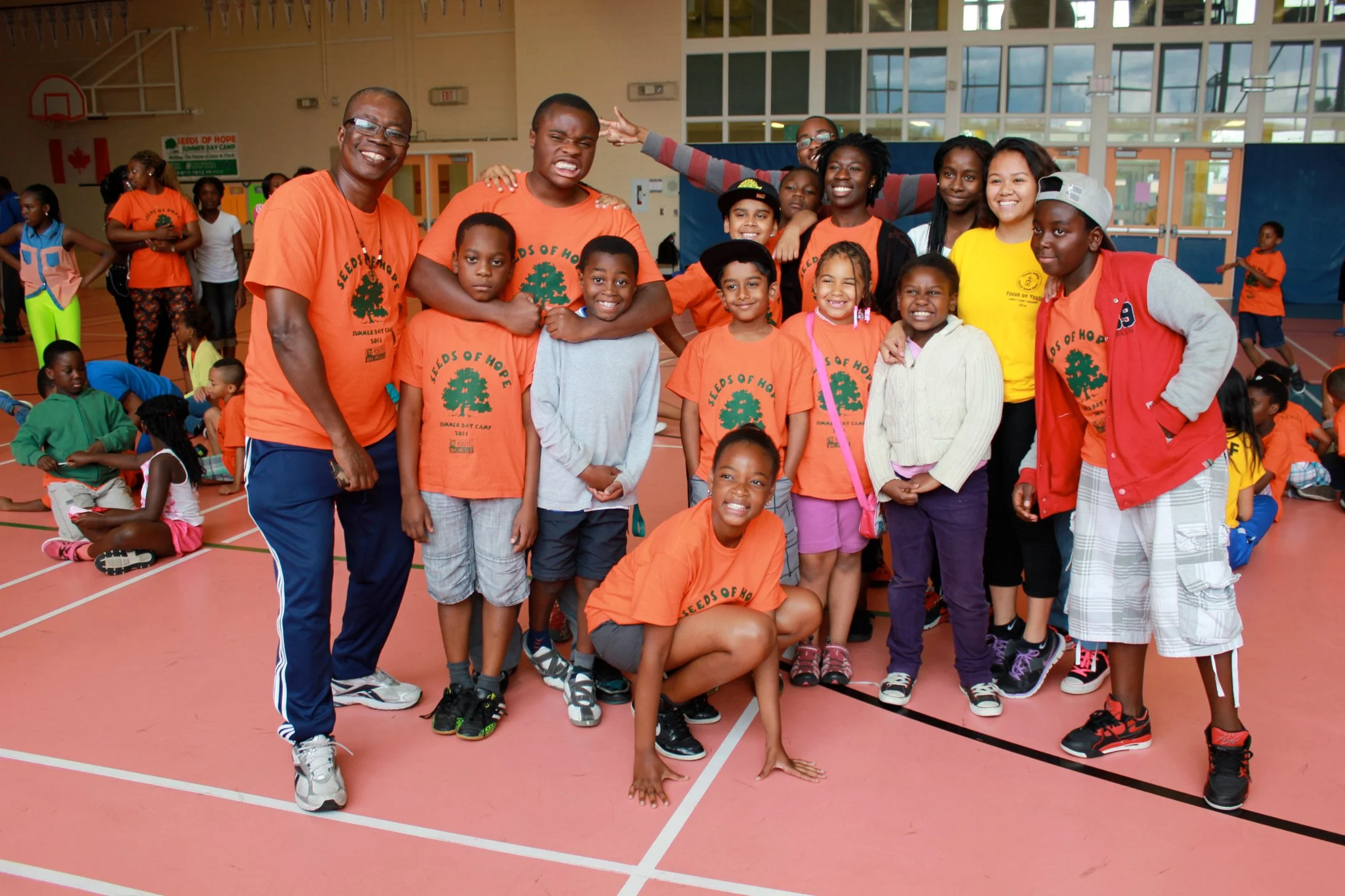 A group of children and adults in a gymnasium, some wearing orange T-shirts with a green tree logo, smiling and posing for a photo.