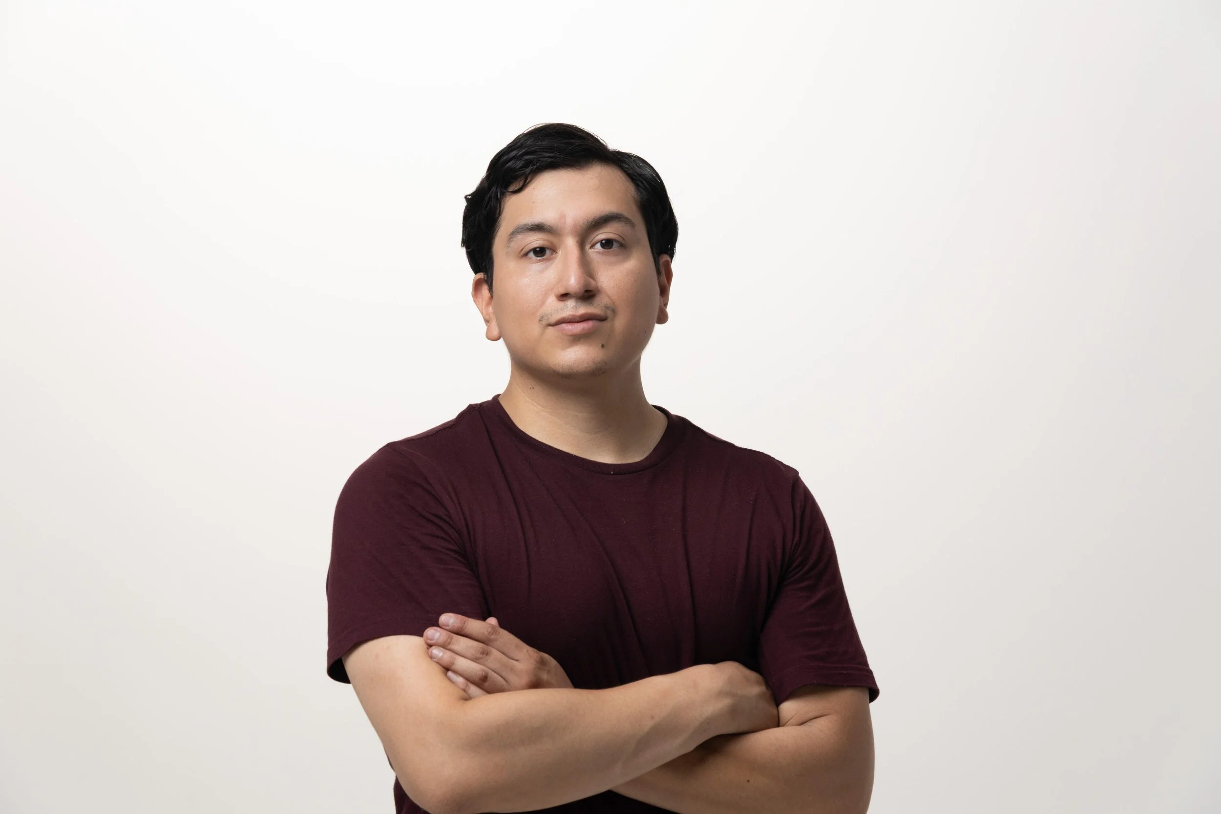 A young man with black hair and light skin, wearing a black T-shirt, standing with arms crossed against a dark background.