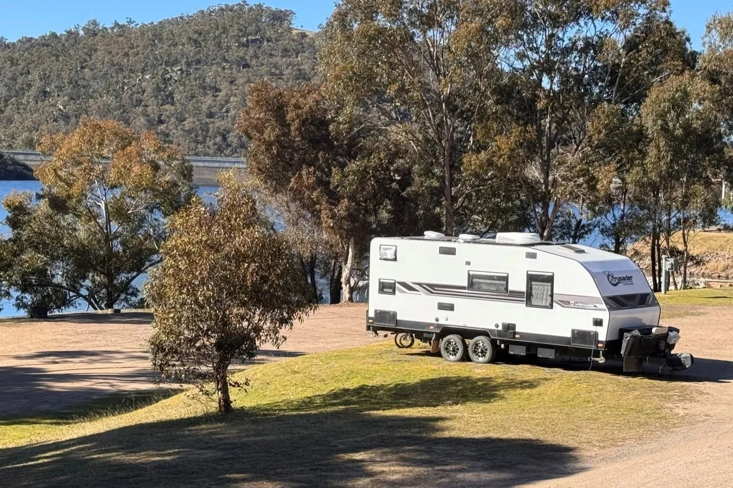A white caravan is parked on a crushed granite pad and green grass surrounded by bushland and the blue Lake Lyell in the background