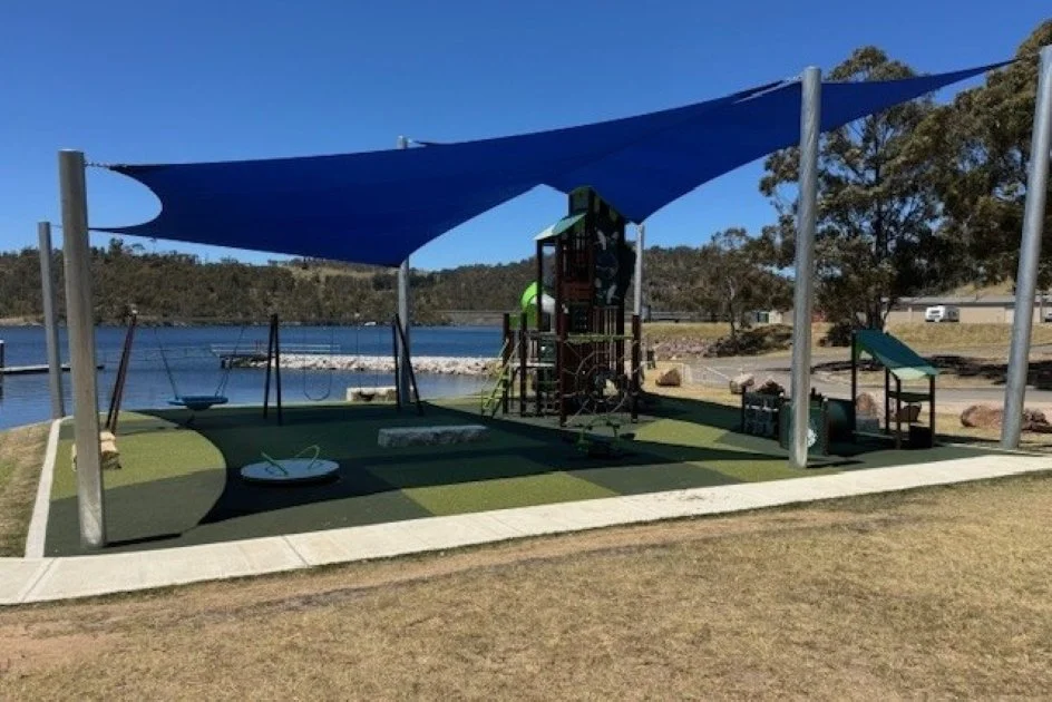Blue sails provide shelter over a green carpeted playground with a tall brown and green tower and slippery slide in the middle