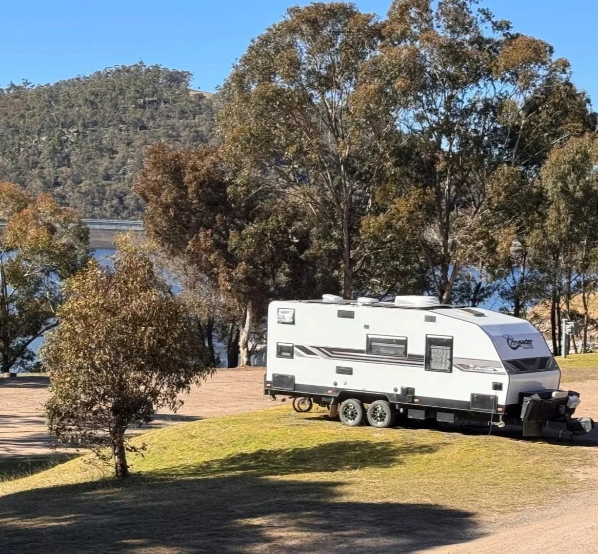 A white caravan with black livery is parked on green grass surrounded by gum trees and a blue lake in the foreground.