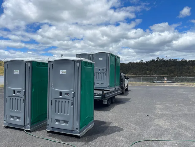 Green and grey portaloos on the back of a ute and on dark grey bituman with a blue lake and bushland in the background
