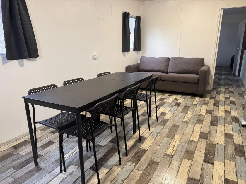 A black table setting with six chairs stands central in the dining room with a grey sofa couch located behind in the lounge room.