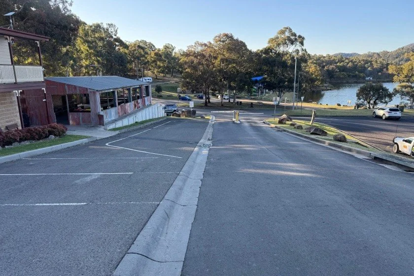 A dark bitumen road leads past a cream two storey building and a red and grey single story building