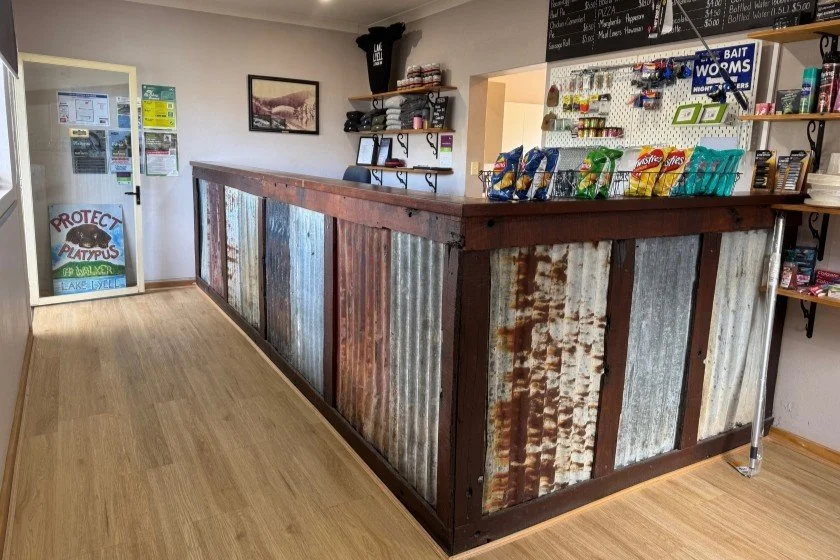 A red, grey and brown corrugated iron clad shop counter is surrounded by yellow-cream timber flooring and shop merchandise behind the counter