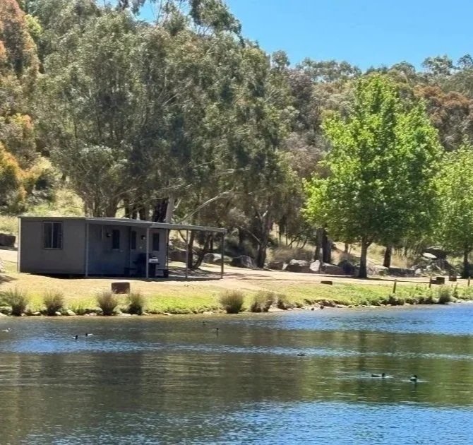 A dark grey cabin sits along the foreshore of a blue lake surrounded by green grass and green gum trees.