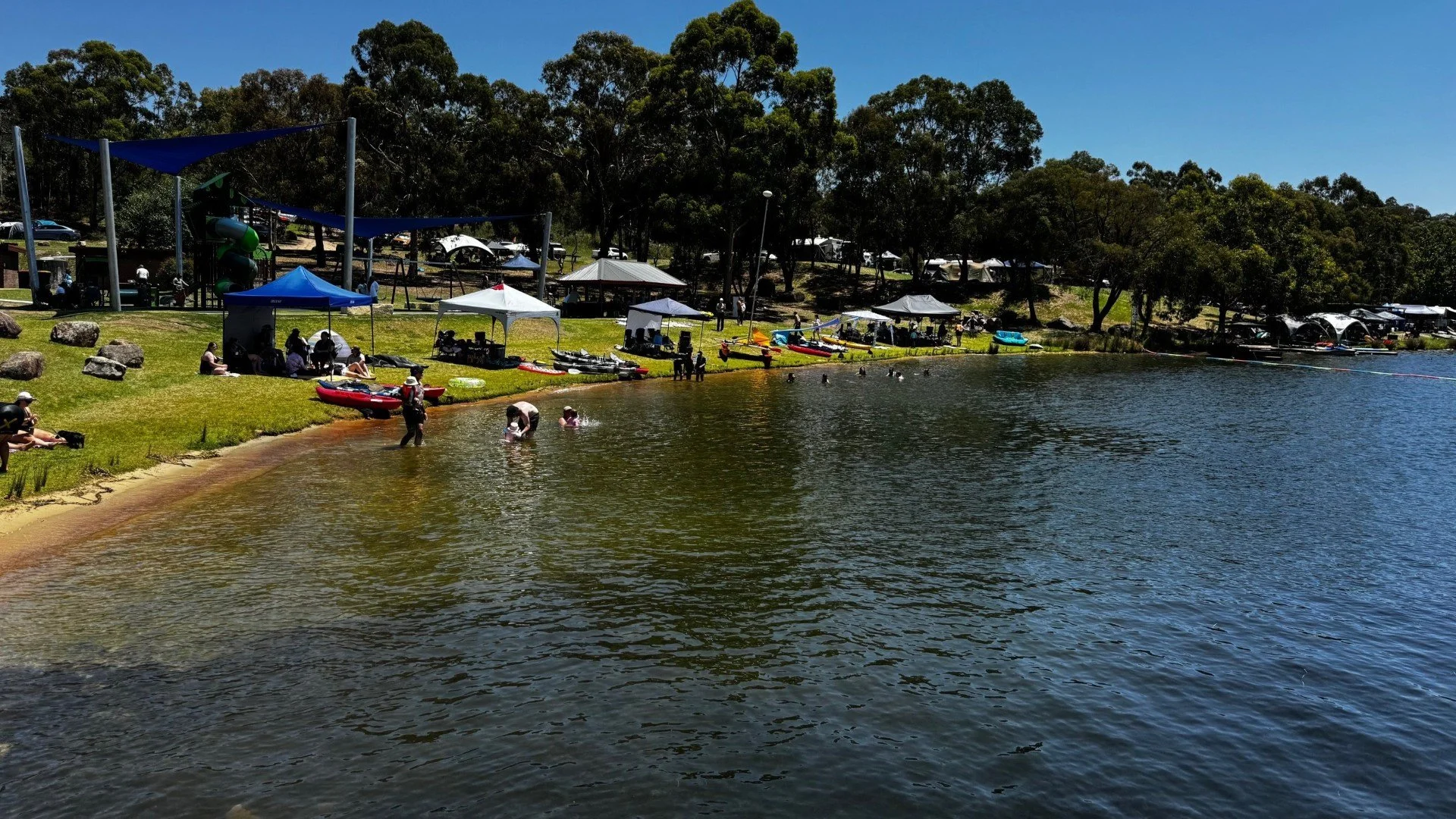 People line the foreshore of a blue lake swimming in the calm water or sitting under the shade of blue and white gazebos