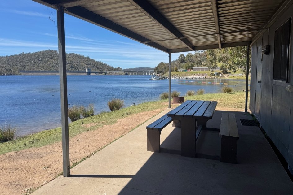 A brown outdoor timber table is sheltered by a grey metal awning looking out over the blue water of Lake Lyell