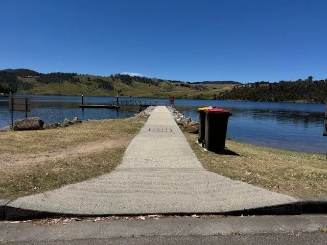 A grey concrete path leads past a yellow and red-lidded bins out to the blue water