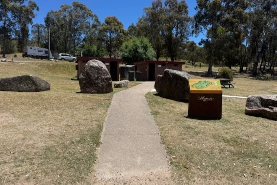 A concrete pathway leads through large rock boulders and past a yellow and red sign to two brown brick buildings in the distance