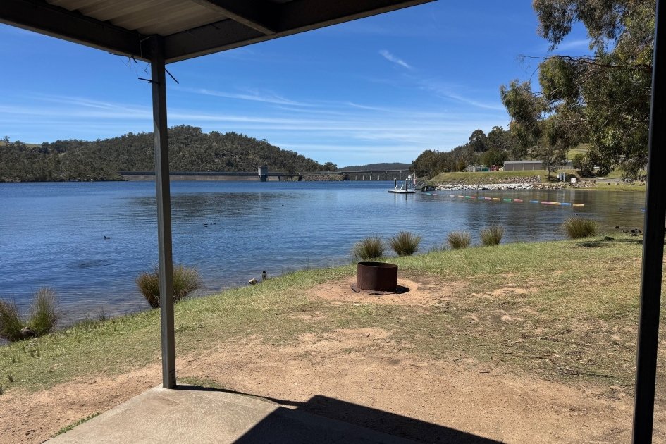 A rusty red fire pit ring is visible along the foreshore from a grey metal awning looking out over the blue water of Lake Lyell