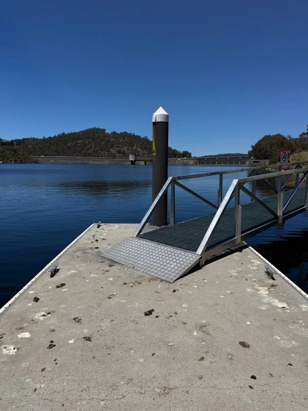 A grey metal ramp with silver metal handrails leads out onto a grey floating pontoon on the blue water