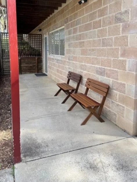 Two brown timber bench seats sit at the front of a cream coloured building with a maroon coloured post to the left