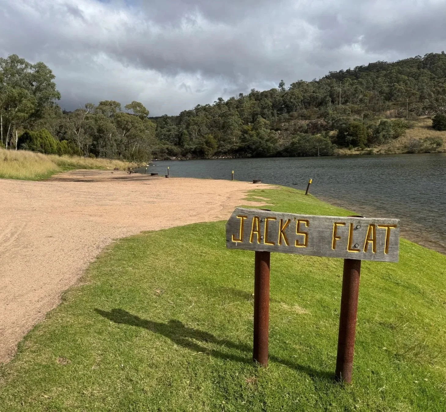 A brown timber sign with yellow writing "Jacks Flat" stands proud at the entrance to bare earth and green grass along the foreshore of a blue lake.