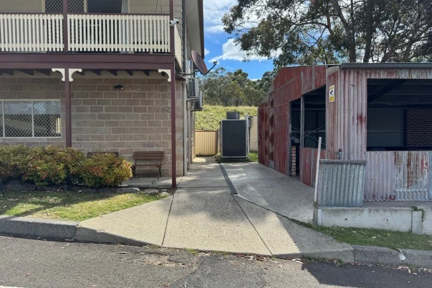 A grey concrete path leads from a dark bitumen road to a cream coloured two-storey brick building