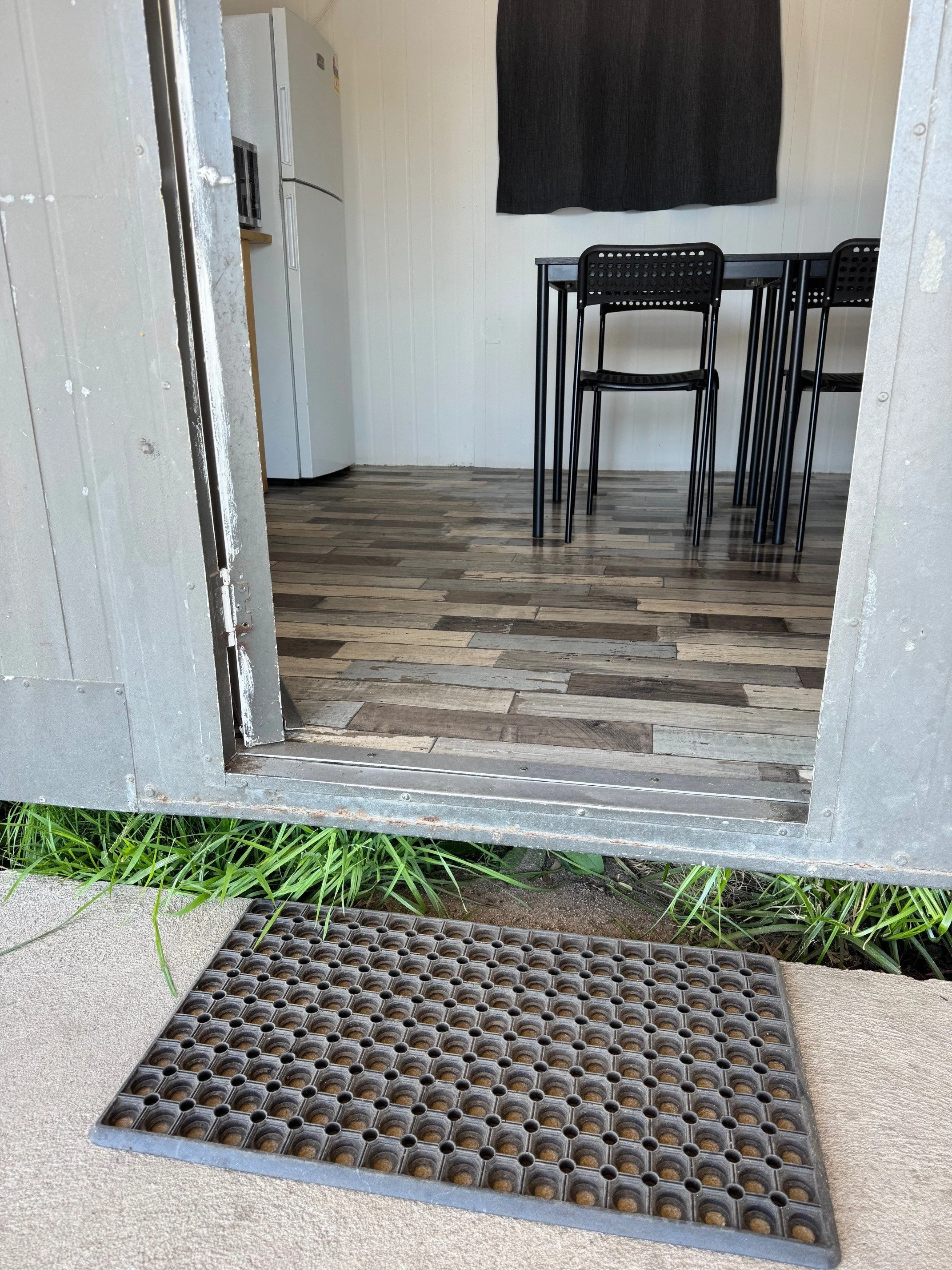 A grey textured rubber mat sits before a step leadingat the entrance into a grey building with a black table setting just inside the door.